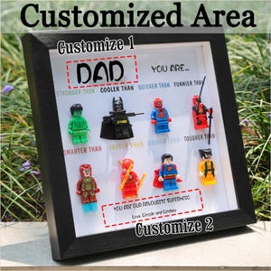 May include: A framed artwork featuring Lego superhero figures. The text "DAD" is displayed above the figures, with phrases like "STRONGER THAN" and "COOLER THAN." The bottom text reads "YOU ARE OUR FAVOURITE SUPERHERO."