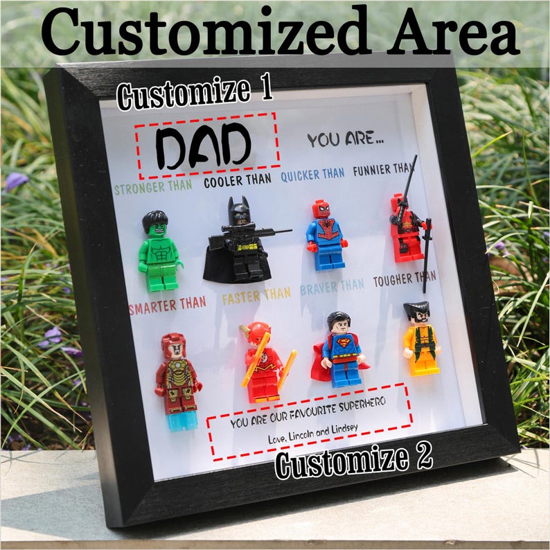 May include: A framed artwork featuring Lego superhero figures. The text "DAD" is displayed above the figures, with phrases like "STRONGER THAN" and "COOLER THAN." The bottom text reads "YOU ARE OUR FAVOURITE SUPERHERO."