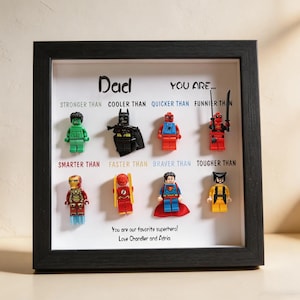 May include: A framed artwork featuring Lego superhero figures. The black frame surrounds a white background with the word "Dad" and phrases like "Stronger Than" and "Cooler Than." The text "You are our favorite superhero! Love Chandler and Adria" is also included.
