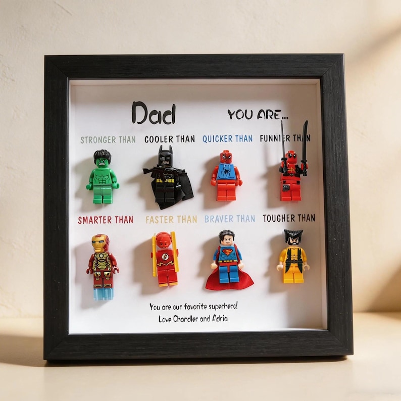 May include: A framed artwork featuring Lego superhero figures. The black frame surrounds a white background with the word "Dad" and phrases like "Stronger Than" and "Cooler Than." The text "You are our favorite superhero! Love Chandler and Adria" is also included.