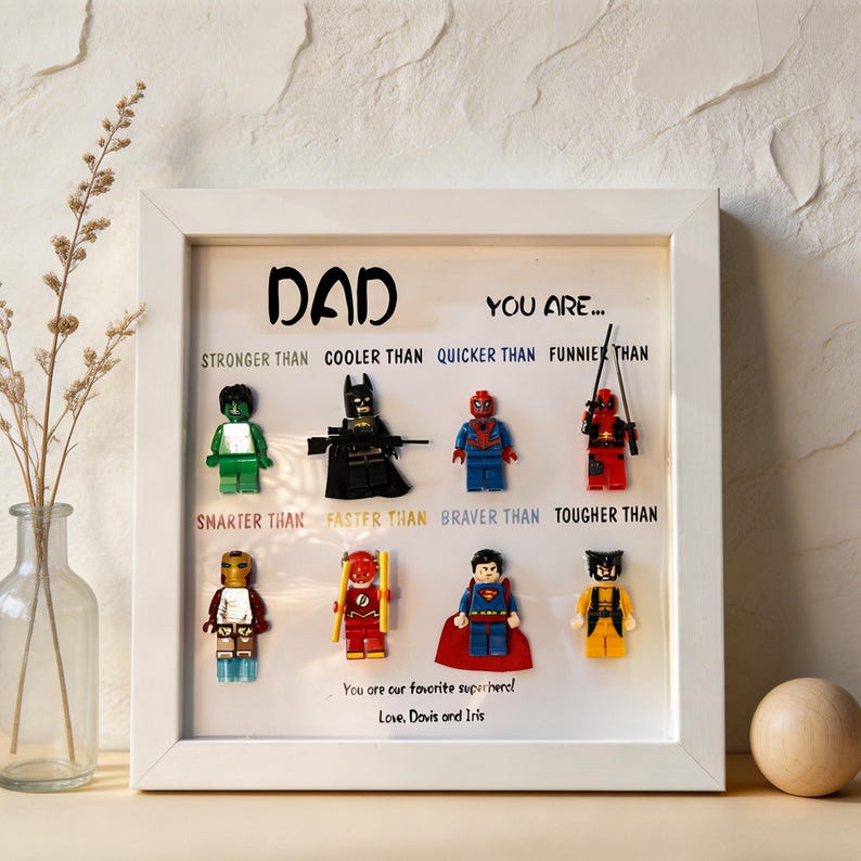 May include: A framed artwork featuring Lego superhero figures. The white frame surrounds a display with the word "DAD" and phrases like "STRONGER THAN" and "BRAVER THAN." The bottom text reads "You are our favorite superhero!"