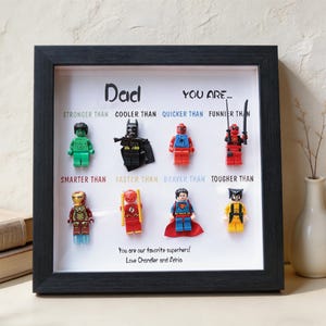 May include: A framed artwork featuring Lego superhero figures. The black frame surrounds a white background with the text "Dad, You are..." and various superhero comparisons. The bottom reads "You are our favorite superhero! Love Chandler and Adria."