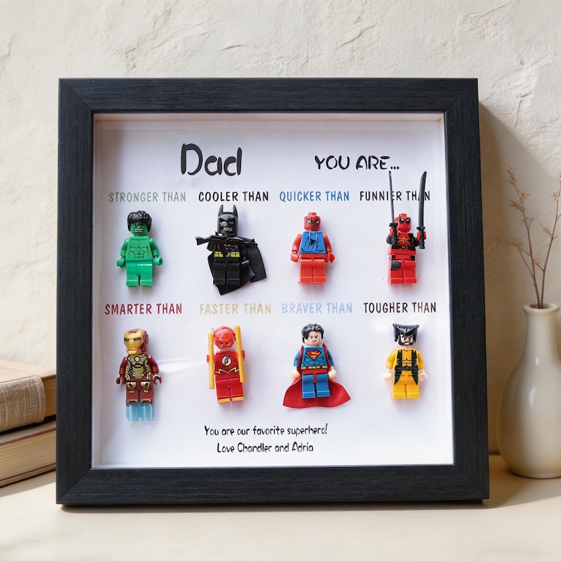 May include: A framed artwork featuring Lego superhero figures. The black frame surrounds a white background with the text "Dad, You are..." and various superhero comparisons. The bottom reads "You are our favorite superhero! Love Chandler and Adria."
