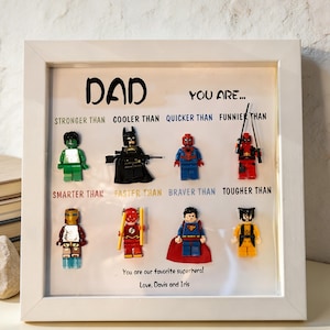 May include: A framed artwork with the word "DAD" and the phrase "YOU ARE..." above Lego superhero figures. The text below the figures reads "STRONGER THAN, COOLER THAN, QUICKER THAN, FUNNIER THAN, SMARTER THAN, FASTER THAN, BRAVER THAN, TOUGHER THAN." The bottom reads "You are our favorite superhero! Love, Davis and Iris."