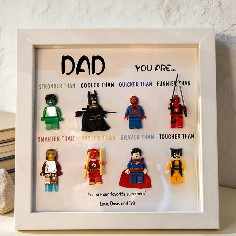 May include: A framed artwork with the word "DAD" and the phrase "YOU ARE..." above Lego superhero figures. The text below the figures reads "STRONGER THAN, COOLER THAN, QUICKER THAN, FUNNIER THAN, SMARTER THAN, FASTER THAN, BRAVER THAN, TOUGHER THAN." The bottom reads "You are our favorite superhero! Love, Davis and Iris."