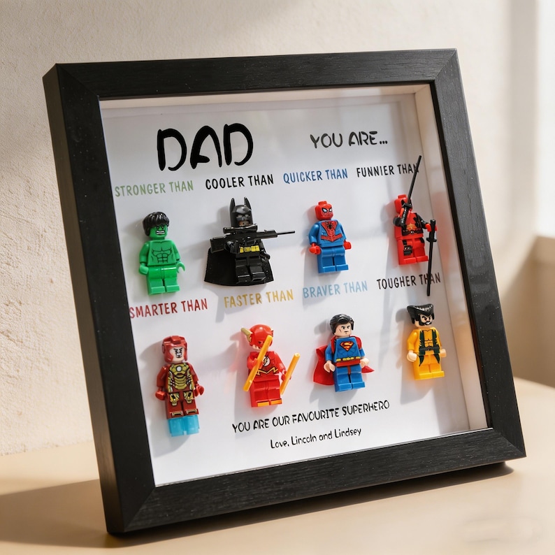 May include: A framed artwork featuring Lego superhero figures. The black frame surrounds a white background with the word "DAD" and phrases like "STRONGER THAN" and "COOLER THAN." The artwork includes Lego versions of superheroes such as Batman, Superman, and Wolverine.