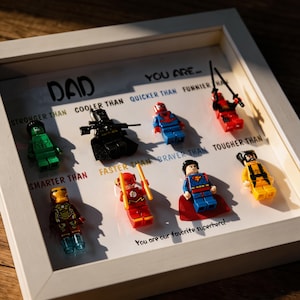 May include: A framed artwork featuring Lego superhero figures. The text reads "DAD, you are..." followed by descriptive comparisons like "STRONGER THAN" and "BRAVER THAN." The bottom text says "You are our favorite superhero!"