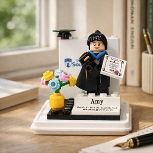 May include: A Lego figurine of a graduate in a black gown and cap, holding a diploma. The figurine is personalized with the name "Amy" and "MA Film & Cultural Management." A small bouquet of flowers in a yellow vase is also included.