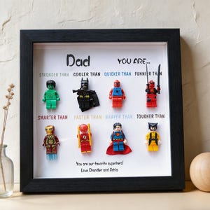 May include: A framed artwork featuring Lego superhero figures. The black frame surrounds a white background with the word "Dad" and phrases like "Stronger Than" and "Braver Than." The text "You are our favorite superhero!" is also included.