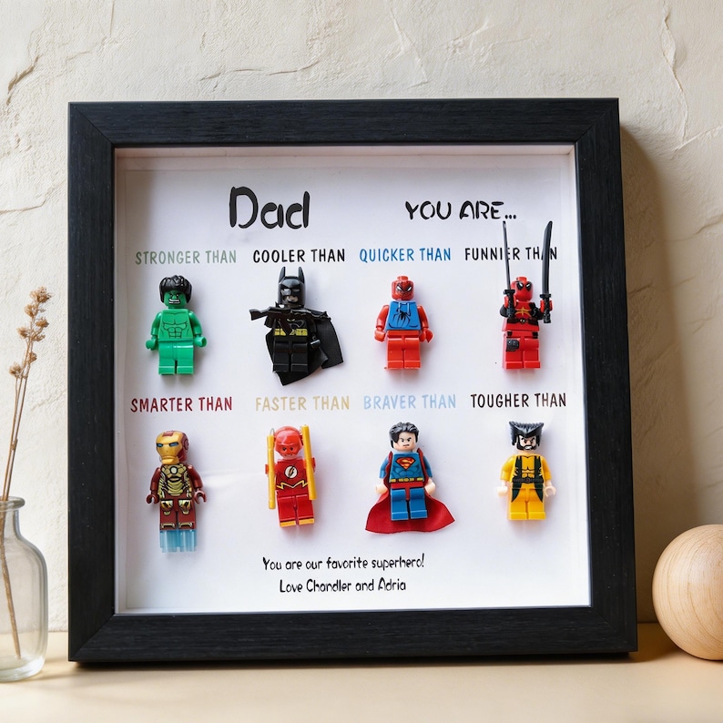 May include: A framed artwork featuring Lego superhero figures. The black frame surrounds a white background with the word "Dad" and phrases like "Stronger Than" and "Braver Than." The text "You are our favorite superhero!" is also included.