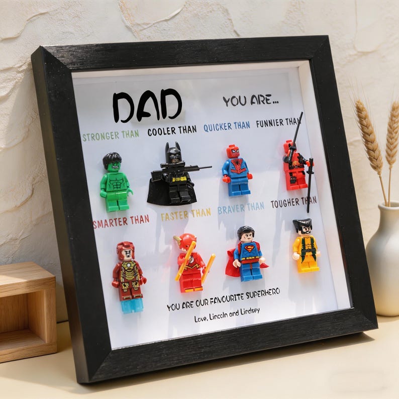 May include: A framed artwork featuring the word "DAD" above various superhero Lego figures. Text below the figures reads "YOU ARE..." and lists qualities like "STRONGER THAN" and "SMARTER THAN." The bottom text says "YOU ARE OUR FAVOURITE SUPERHERO."
