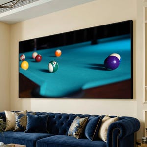 May include: A large framed print of a pool table with several billiard balls on a teal felt surface. The balls are various colors, including yellow, green, red, and purple. The print is displayed above a blue sofa with decorative pillows.