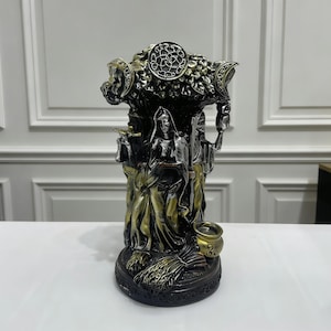 May include: A detailed statue featuring three figures, with a central female figure holding a staff. The statue has a dark bronze and silver finish, with intricate details on the figures and the base. The top features a circular design.