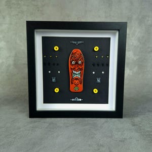 May include: Framed skateboard art. An orange skateboard deck with a red, angry face graphic is mounted on a black background. The artwork includes hardware and yellow wheels, and is encased in a black frame.