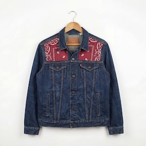Può includere: Giacca di jeans blu scuro con motivi paisley rossi sulle spalle. La giacca ha un design classico con bottoni, due tasche sul petto e una piccola etichetta rossa. Appesa a una gruccia di legno.