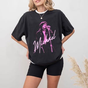May include: Black t-shirt with a pink graphic of a singer with a microphone, and the name "Michael" in cursive. The shirt has a crew neck and short sleeves. The model is wearing black shorts.