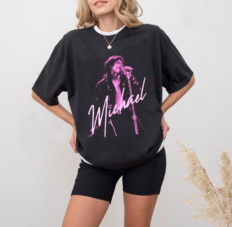 May include: Black t-shirt with a pink graphic of a singer with a microphone, and the name "Michael" in cursive. The shirt has a crew neck and short sleeves. The model is wearing black shorts.