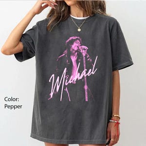 May include: A pepper-coloured t-shirt with a pink graphic of a singer on stage with a microphone. The singer is wearing a jacket and the name "Michael" is written in pink script below the image. The shirt has short sleeves.
