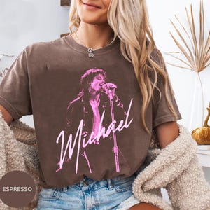 May include: Espresso-coloured t-shirt featuring a pink graphic of a singer with a microphone, with the name "Michael" in a cursive font. The shirt has a vintage, worn look.