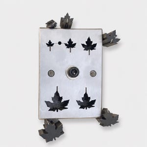May include: A rectangular brushed metal plate featuring five laser-cut maple leaf shapes of varying sizes. The plate includes a central black bolt and two smaller circular fasteners. Four additional dark metal maple leaf shapes are attached to the corners.
