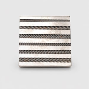 May include: A square metal block featuring six horizontal rows of embossed rope and braided patterns. The surface has a metallic silver finish with dark, recessed detailing within the textured lines, creating a distinct contrast across the face.