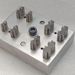 Può includere: Un utensile rettangolare in metallo color argento con diversi componenti dentati in rilievo, distanziati uniformemente. È visibile un bullone esagonale centrale di colore scuro. L'utensile è progettato per lavori di precisione, probabilmente in ambito manifatturiero o ingegneristico.