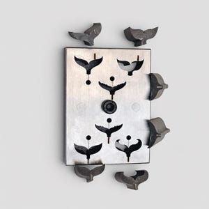 May include: A rectangular silver-toned metal plate featuring six cutouts in the shape of whale tails, with a central black circular knob. Several detached, three-dimensional dark metal whale tail components are arranged around the plate.