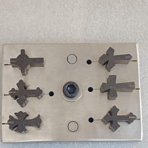 Puede incluir: Una placa metálica con seis objetos en forma de cruz de color gris oscuro dispuestos en dos filas. La placa tiene un elemento circular central y varias hendiduras circulares más pequeñas. Las cruces son de diseños variados.