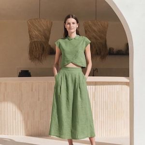 May include: A sage green two-piece set consisting of a sleeveless cropped top with a high neckline and side button detailing, paired with a matching high-waisted midi skirt featuring a pleated front design and side pockets.