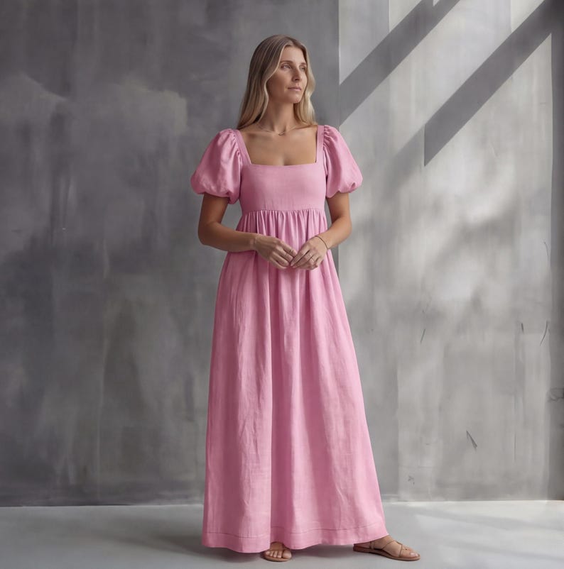 May include: A floor-length pink dress featuring short puff sleeves, a square neckline, and a fitted bodice that transitions into a loose, flowing skirt. The garment is constructed with a simple, clean design and a solid light pink color throughout.