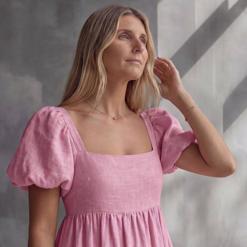 May include: A light pink dress featuring a square neckline and short puff sleeves. The garment is crafted from a textured fabric and includes a gathered waistline that flows into a full skirt. A thin gold necklace and a matching gold bracelet are worn.