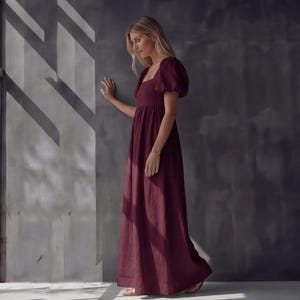 May include: A burgundy maxi dress with a square neckline and short, puffed sleeves. The dress has a fitted bodice and a flowing skirt. The woman is wearing a gold bracelet and sandals. The background is a gray wall with shadows.