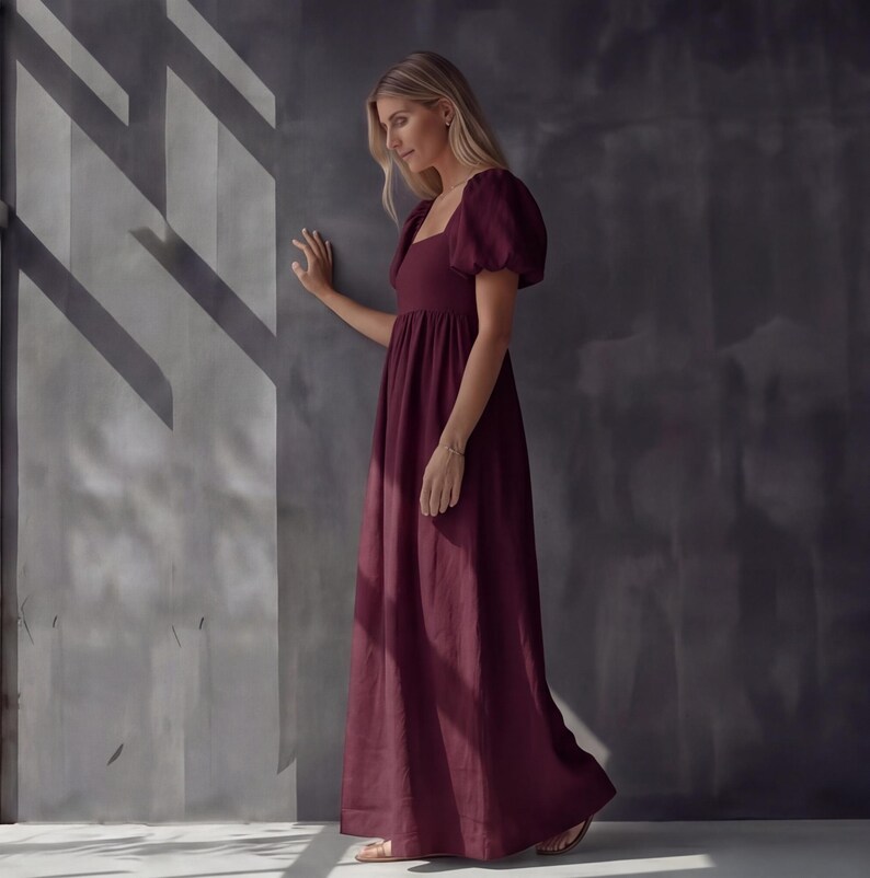 May include: A burgundy maxi dress with a square neckline and short, puffed sleeves. The dress has a fitted bodice and a flowing skirt. The woman is wearing a gold bracelet and sandals. The background is a gray wall with shadows.