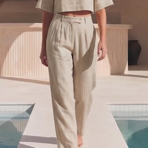 May include: High-waisted trousers in a light beige color featuring a straight-leg cut, front pleats, a waistband with belt loops, and a button and zipper closure. The trousers are paired with a matching short-sleeved cropped top.
