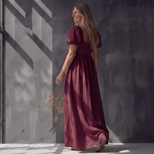 May include: A burgundy maxi dress with short, puffed sleeves and a square neckline. The dress has a fitted bodice and a flowing skirt. The model is wearing the dress and walking away from the camera. The dress is made of a soft, flowing fabric.