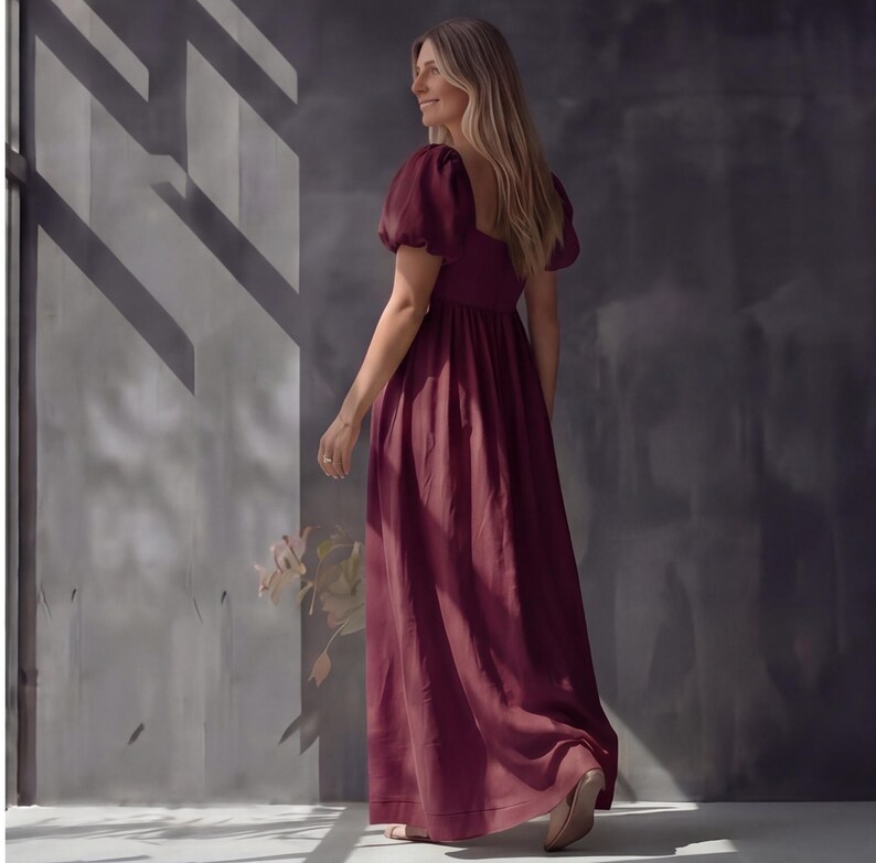May include: A burgundy maxi dress with short, puffed sleeves and a square neckline. The dress has a fitted bodice and a flowing skirt. The model is wearing the dress and walking away from the camera. The dress is made of a soft, flowing fabric.
