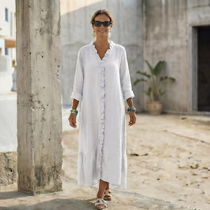 May include: A white, long-sleeved dress with a ruffled trim down the front and a tiered hem. The dress has a V-neck and is worn with sunglasses and sandals. The dress appears to be made of a lightweight fabric.