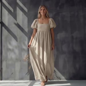 May include: A beige linen maxi dress with a square neckline and short, puffed sleeves. The dress has a gathered waist and a flowing skirt. The model is standing in front of a gray wall with shadows.