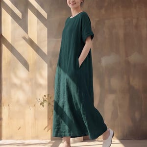 May include: A dark forest green maxi dress featuring a round neckline, short sleeves with folded cuffs, and side pockets. The garment has a loose, straight-cut silhouette that falls to the ankles, crafted from a solid-coloured, opaque fabric.