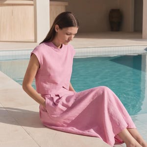 May include: A two-piece set consisting of a light pink sleeveless top with a high neckline and a matching light pink midi skirt. The top features a cropped length with a straight hem, while the skirt has a high waist and side pockets.