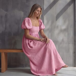 May include: A floor-length pink dress featuring short puff sleeves, a square neckline, and a gathered bodice. The garment has a loose, flowing skirt that extends to the ankles, crafted from a solid-colored fabric with a visible texture.