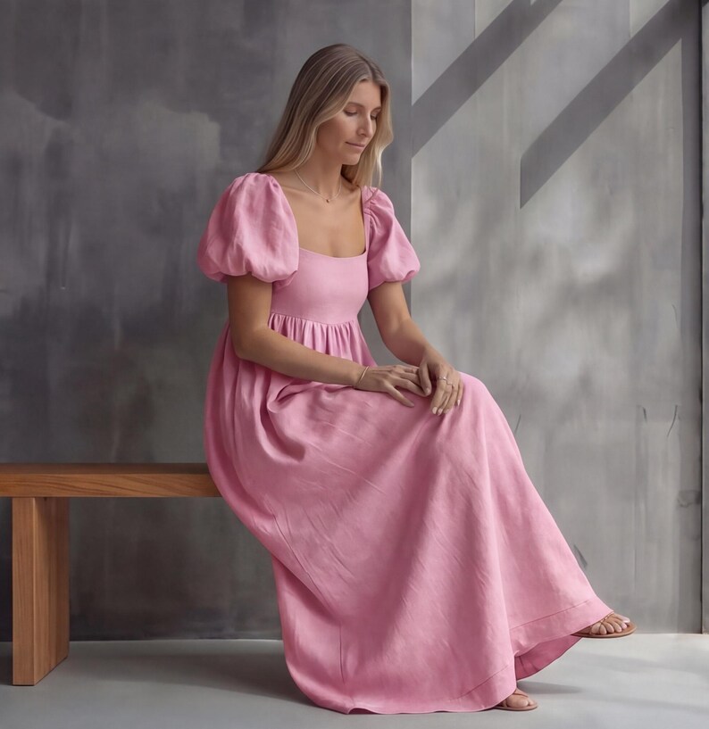 May include: A floor-length pink dress featuring short puff sleeves, a square neckline, and a gathered bodice. The garment has a loose, flowing skirt that extends to the ankles, crafted from a solid-colored fabric with a visible texture.