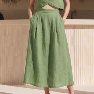May include: High-waisted sage green wide-leg trousers featuring a structured waistband, front pleats, and side pockets. The garment is crafted from a textured fabric and falls to a midi length, offering a loose, flowing silhouette with a clean finish.
