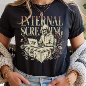 May include: Black t-shirt with the words "INTERNAL SCREAMING" in a distressed font. The graphic features a skeleton wearing a hoodie, sitting at a desk with a laptop displaying "ERROR 404", surrounded by floral elements.