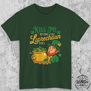 May include: Green t-shirt featuring a cartoon leprechaun with a pot of gold and the text "Kiss Me Before The Leprechaun Does." The design includes shamrocks and gold coins, perfect for St. Patrick's Day.