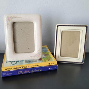 May include: Two decorative picture frames. One is off-white with a floral design and a crackle finish. The other is off-white with a navy blue and gold border. The frames are resting on top of two books with the titles "Love Comes Softly" and "My Fairy Godmother".