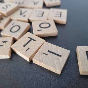 May include: Wooden Scrabble tiles with black letters and numbers are scattered on a dark surface. The letters visible include "T", "O", "E", "I", and "Y". The tiles are made of light-colored wood.
