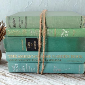 May include: A stack of five vintage books in shades of green and teal, bound together with twine. The book titles are visible, including "Shelter" and "The American Home Garden Book". The books are arranged on a light-colored wooden surface.