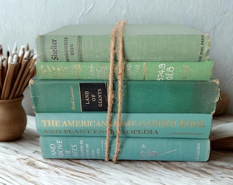 Puede incluir: Una pila de cinco libros antiguos en tonos verdes y turquesas, atados con cuerda. Los títulos, como "Shelter" y "The American Home Garden Book", son visibles. Los libros están dispuestos sobre una superficie de madera clara.