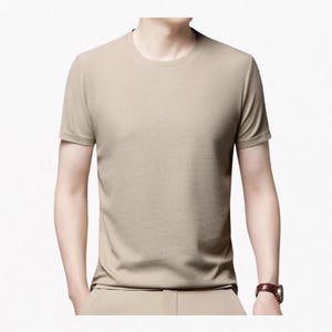 Könnte beinhalten: Ein kurzärmeliges T-Shirt in einfarbigem Beige mit einem fein strukturierten Gittermuster auf dem gesamten Stoff. Das Kleidungsstück verfügt über einen klassischen Rundhalsausschnitt und einen geraden Saum, was für ein sauberes und einheitliches Erscheinungsbild sorgt.